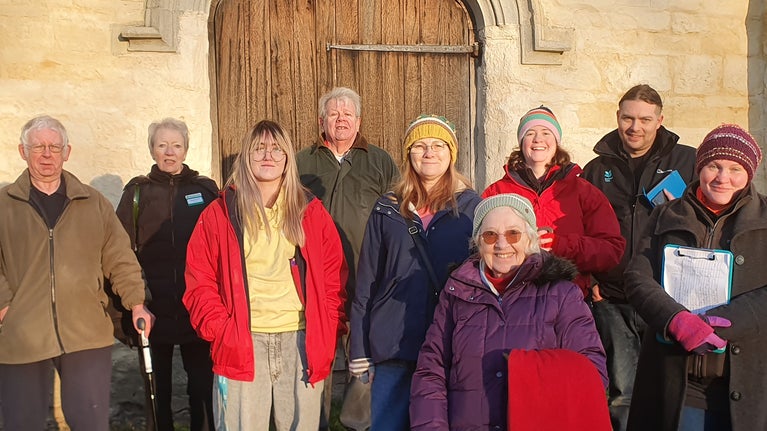 A group of 9 people of various ages, stood together smiling, in front of a some old stonework and a big wooden door, most are wearing winter coats, and the evening light is glowing gold across their faces and the stonework behind them